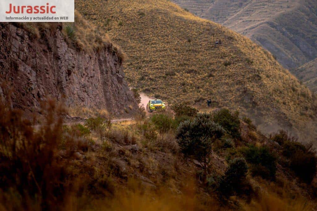 A rally truck speeds through a rugged mountainous path under a clear sky.